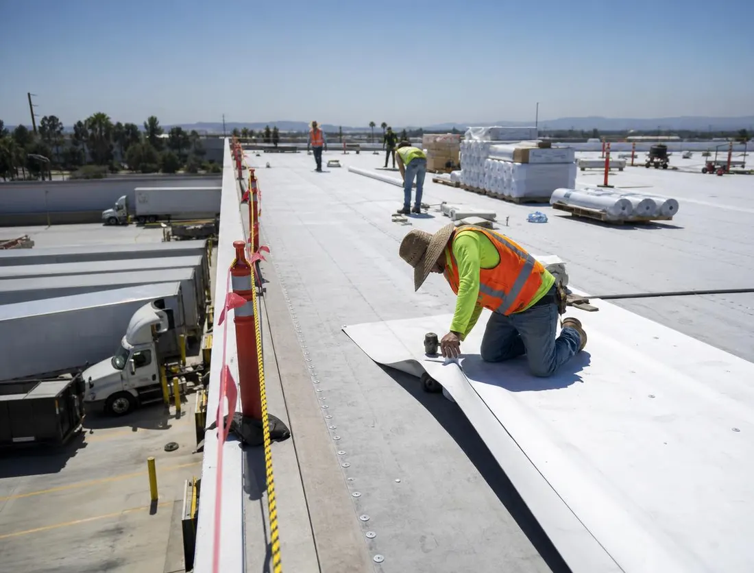 Roofing crew working a taped-off zone on a warehouse roof while trucks continue loading below
