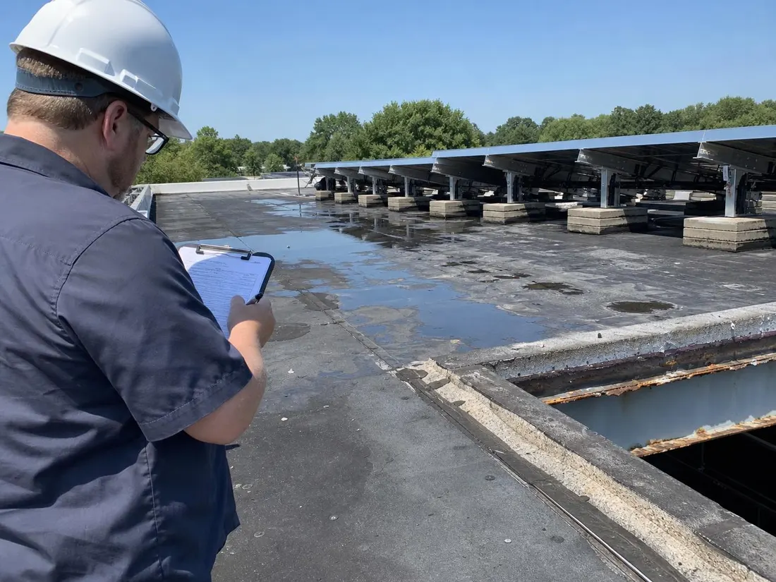 Engineer inspecting an aging flat roof with ponding water beside a ballasted solar mount